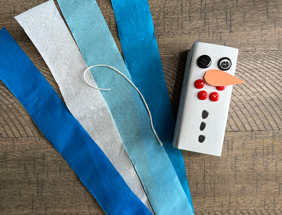 white construction paper is put over the juice juice box to create the white snowman. buttons are used for the eyes and mouth, a carrot shaped nose is cut out from the orange construction paper, and the black marker is used for details such as the snowmans buttons.