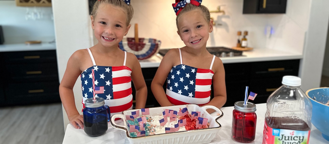 Two children in american flag outfits in front of a tray with rice crispy treats and mason jars with blue and red juice
