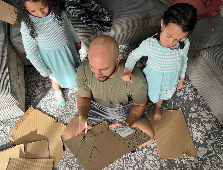 A man and two children sit on the floor surrounded by cardboard boxes. The man is cutting out a sweater shape from cardboard box